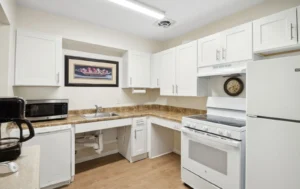 A clean, white kitchen with brown countertops, white appliances, a coffee maker, and microwave. A wall clock and framed art hang above the sink area with visible plumbing.