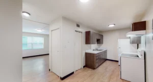 Unfurnished apartment interior showing a kitchen with brown cabinets, white appliances, and a view into a bright living area with wood-style flooring.