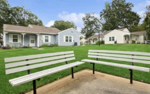 Two white benches face each other on a concrete pad with green grass and single-story houses in the background under a partly cloudy sky.