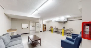 A brightly lit office lounge with sofas, chairs, a coffee table, workstations, a bookshelf, and a fire extinguisher mounted on the wall.