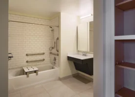 Accessible bathroom with a bathtub featuring grab bars and a transfer bench, a wall-mounted sink, a mirror, and tiled floors and walls.