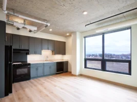 Modern apartment kitchen with dark cabinets, black appliances, light wood flooring, large window, and exposed concrete ceiling with visible piping.