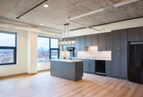 Modern kitchen with gray cabinets, black appliances, a light wood island, pendant lights, and large windows overlooking a cityscape.