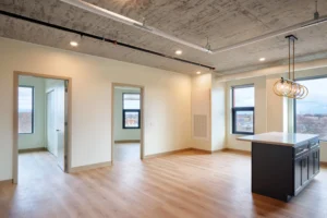 Modern empty apartment with light wood floors, exposed concrete ceiling, large windows, and a kitchen island with pendant lights. Two open doorways lead to separate rooms.