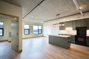 Modern, unfurnished apartment with open-plan kitchen and living area, light wood floors, large windows, and exposed concrete ceiling.