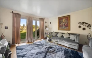 A living room with a glass coffee table, gray sofa with cushions, patterned rug, wall art, and sliding glass doors with curtains opening to a balcony and green outdoor area.