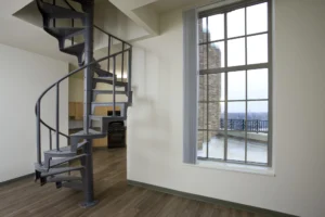A metal spiral staircase next to a large window in a modern apartment with wood flooring, white walls, and an outdoor balcony visible.