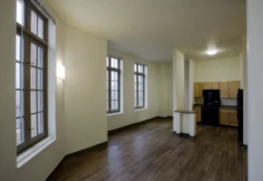Unfurnished apartment with large windows, beige walls, wood-style flooring, and an open kitchen featuring light wood cabinets and black appliances.