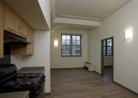 Small apartment kitchen with an electric stove, wood cabinets, and adjacent living area featuring a window, radiator, and hardwood floors. Walls are painted white with minimal decor.