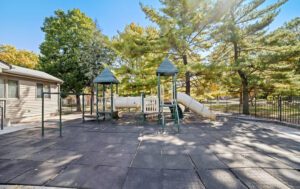 Outdoor playground with two small play structures connected by a covered tunnel slide, surrounded by trees and a black metal fence. Rubber tiles cover the ground.