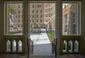 View through an archway showing a walkway, landscaped courtyard, and a multi-story brick apartment building in the background.