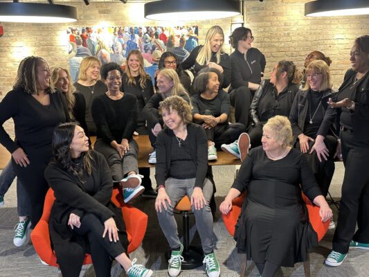 A group of fourteen women, all dressed in black outfits and green sneakers, sit and stand together indoors, smiling and conversing in a casual setting.