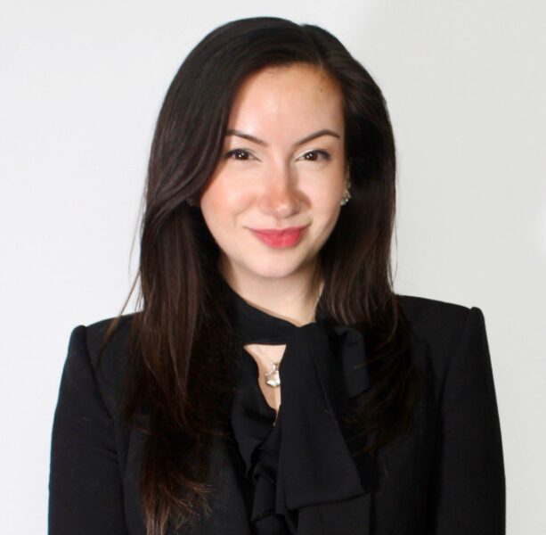 A woman with long dark hair, wearing a black blazer and a black blouse with a bow, stands against a plain white background, smiling slightly at the camera.