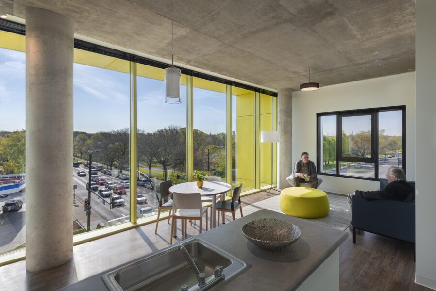 Two people sit and talk in a modern, sunlit office lounge with large windows overlooking a city street and trees. A table, chairs, and a yellow ottoman are in the room.