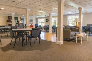 Spacious lounge area with tables, chairs, sofas, large windows, and natural light. A lamp sits on a side table near the seating area.