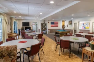 A recreation room with round tables and red chairs, a pool table, an American flag on the wall, and a bulletin board in the background.