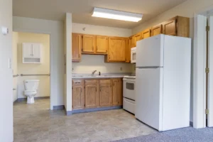 Small kitchen with wooden cabinets, white appliances including a refrigerator and stove, and a view into a bathroom with a toilet in the background.