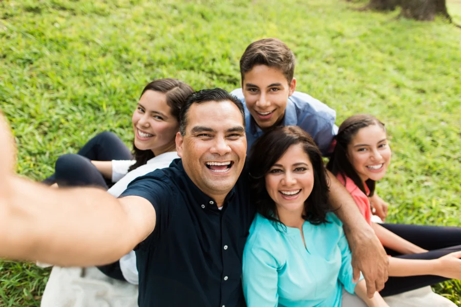 A group of five people, consisting of two adults and three teenagers, sit on grass, smiling at the camera as one person takes a selfie.