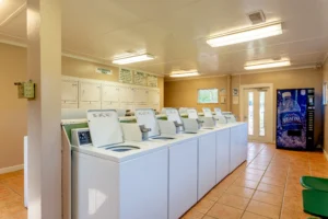 A clean laundry room with a row of white washing machines, stacked dryers against the wall, and a blue Aquafina vending machine near the door.