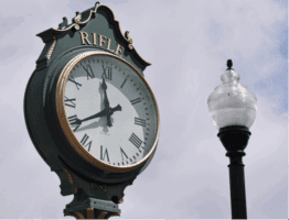 A green street clock with Roman numerals showing 11:02 stands next to a lamp post. The clock has "RIFLE" and "1882" written on it.