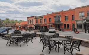 Outdoor metal tables and chairs are set up on a sidewalk patio facing a quiet street with parked cars and brick storefronts under a cloudy sky.