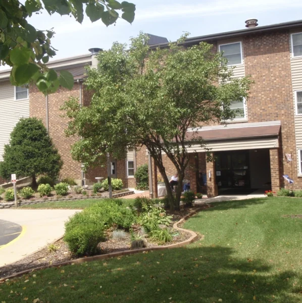 A three-story brick apartment building with landscaped gardens and a sidewalk leading to the main entrance, partially shaded by trees.