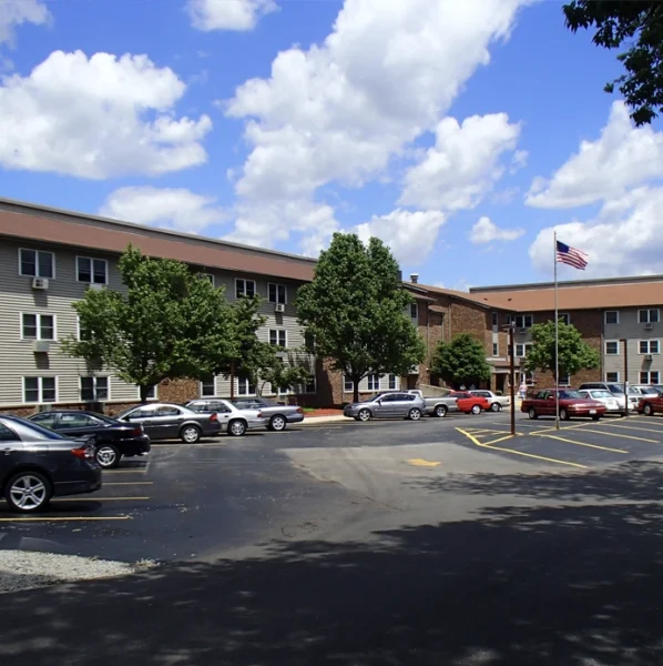 Three-story apartment building with parked cars in front, green trees along the sidewalk, and an American flag on a flagpole, under a partly cloudy sky.
