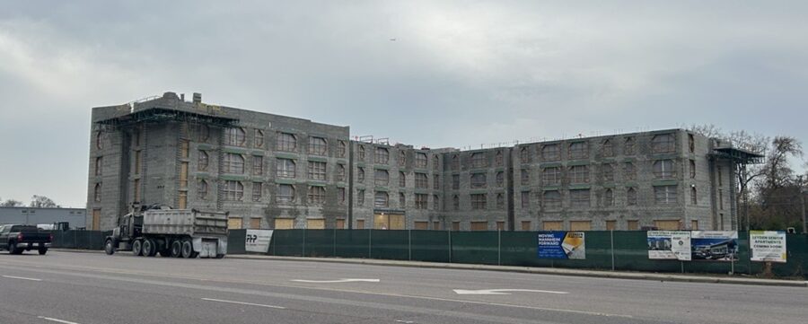 A four-story building under construction is surrounded by green fencing along a roadway, with a dump truck parked in front and various construction signs visible.