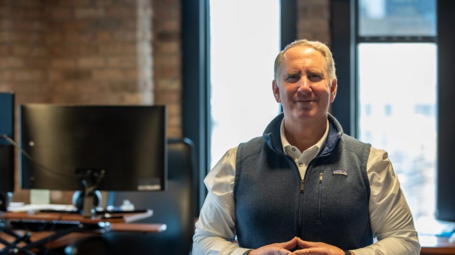 A man wearing a blue vest and white shirt stands with hands clasped in an office with brick walls, windows, and computer monitors in the background.