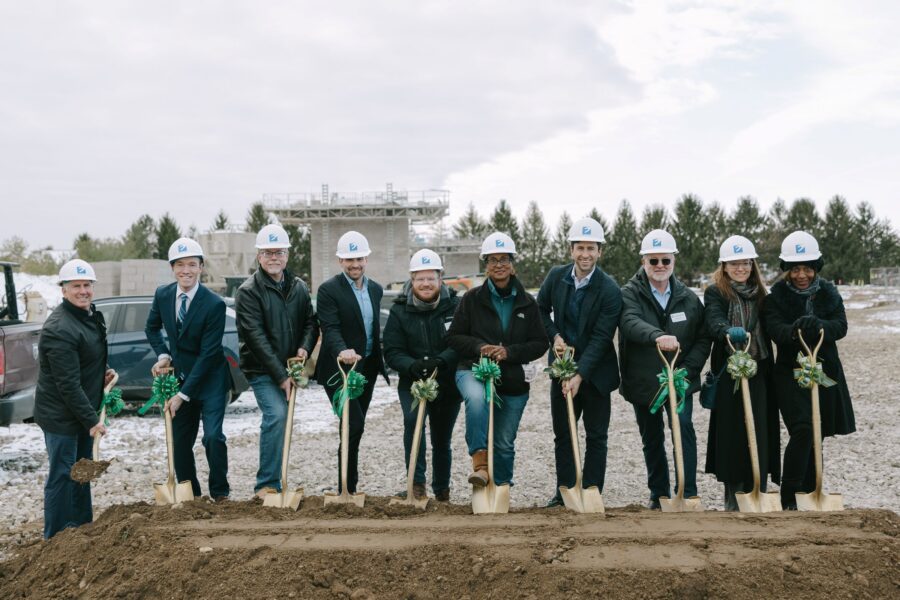 Nine people wearing hard hats and holding shovels stand in a row at a groundbreaking ceremony on a construction site.
