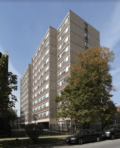 A mid-rise, beige and brown apartment building with many windows, set behind a fence with cars parked along the street and trees in the foreground.