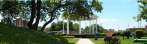 A white gazebo stands in the center of a landscaped park with green trees, bushes, flowers, and blue trash bins on a sunny day.