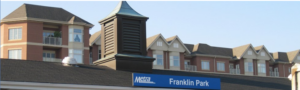 A Metra station sign reading "Franklin Park" is in the foreground, with apartment buildings visible in the background.