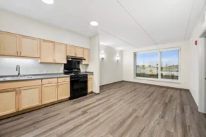 Empty apartment room with wood-style flooring, light wood cabinets, black stove, and a large window facing outside.