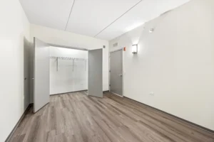 Empty room with light-colored walls, wood-style flooring, an open closet with shelves, and a closed door on the left. Ceiling has visible seams and a wall-mounted light fixture.