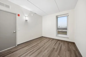Unfurnished room with a gray door, white walls, a wall-mounted light, wood-style flooring, and a large window with an outdoor view.