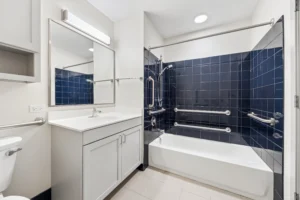 Accessible bathroom with a blue-tiled bathtub, multiple grab bars, a shower chair, a white vanity sink, large mirror, and toilet in a well-lit space.