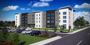 A modern four-story apartment building with a parking lot in front, several parked cars, and a few people walking near landscaped green areas.