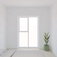Minimalist bedroom with a large window, white walls, a bed in the foreground, and a green potted plant in the corner.