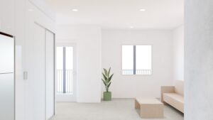 Minimalist living room with white walls, a beige sofa, wooden coffee table, potted plant, and large windows letting in natural light.