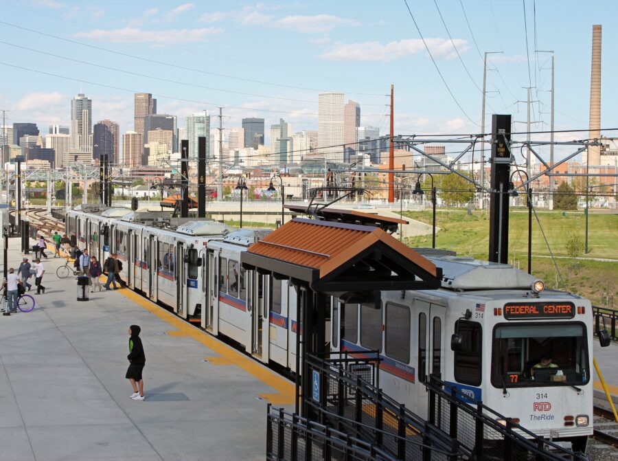 A light rail train at a station platform with people waiting, set against a city skyline in the background on a clear day.