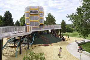 Children play on a modern playground featuring a large hexagonal climbing structure, slides, and tunnels, surrounded by trees and grass under a partly cloudy sky.