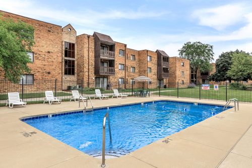 Rectangular outdoor swimming pool with metal handrails, surrounded by lounge chairs and a fence, in front of a multi-story brick apartment building.