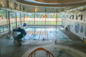 Indoor swimming pool with lap lanes, water slide, and large windows; some people are swimming, and colorful decorations hang near the ceiling.