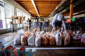 Volunteers wearing gloves pack grocery bags with food items at a community pantry. Two people work in the background, while a person in the foreground organizes bags of groceries on the floor.