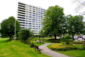 A multi-story apartment building stands behind a landscaped park with benches, trees, yellow flowers, and a parking lot on the right side.