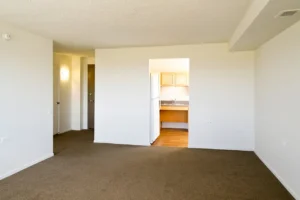 Empty apartment room with beige carpet, white walls, and an open doorway leading to a kitchen with wooden cabinets and a refrigerator. Light fixture and door visible in the hallway.