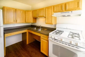 Small kitchen with light wood cabinets, white gas stove, double sink, refrigerator, dark countertops, and wood flooring.