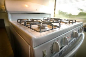 White gas stove with four burners and control knobs, positioned next to a window with blinds partially open.