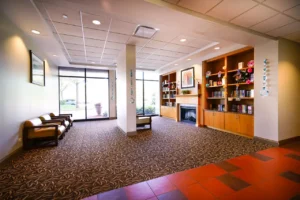 A waiting room with chairs on the left, a bookshelf and fireplace on the right, patterned carpet, and large windows letting in natural light.